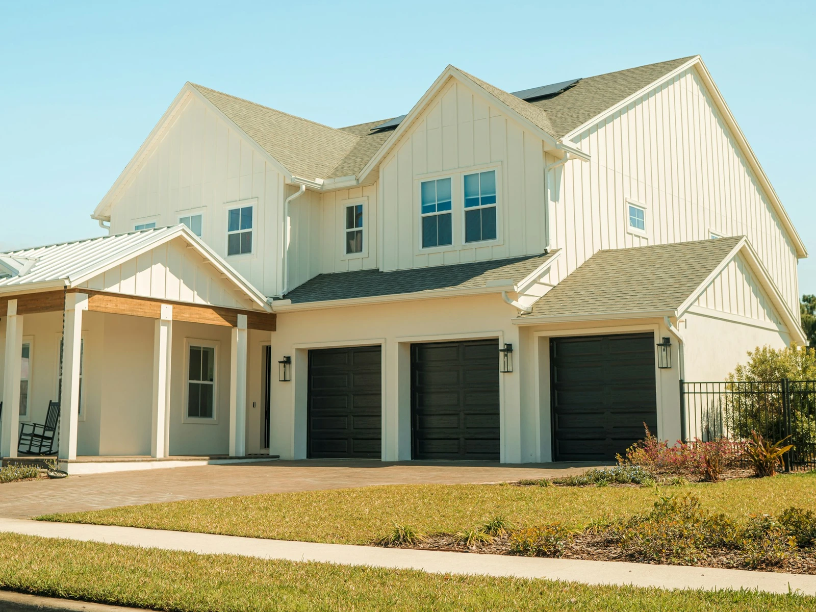 A white board-and-batten home with gabled roofs, three dark garage doors, black lanterns and a green lawn.