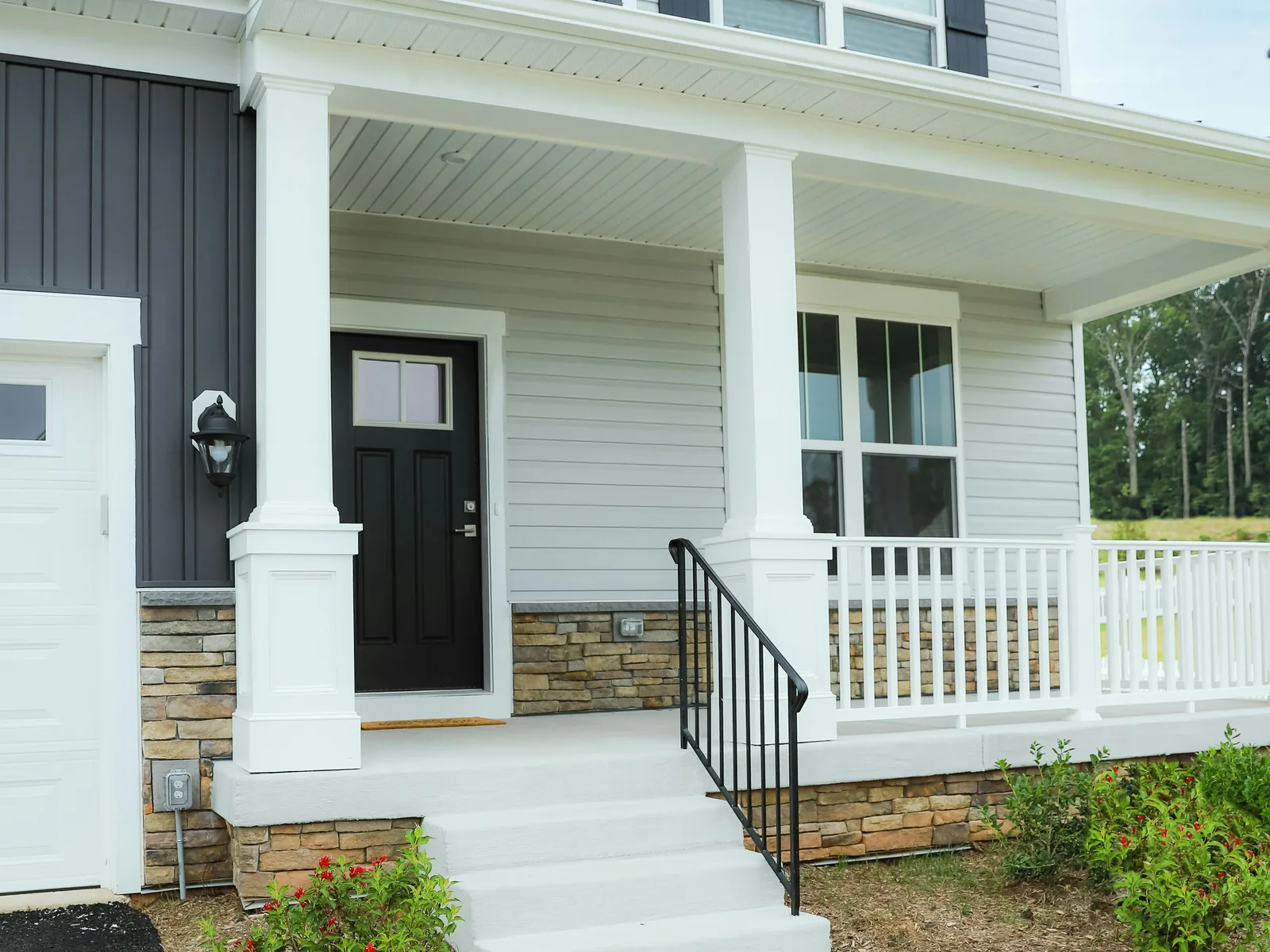A white and charcoal house entrance with a covered porch, square white columns, a black front door, stone veneer bases, white railings and a concrete walk.