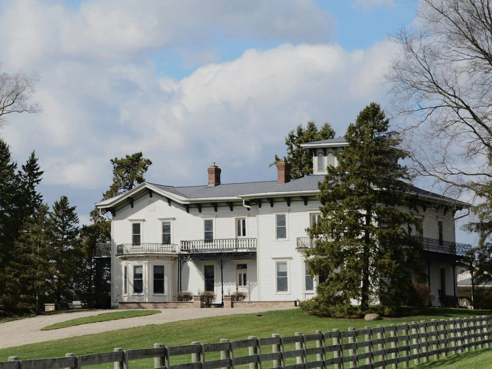 A white country estate with tall sash windows, mature trees, a green lawn and dark timber fencing.