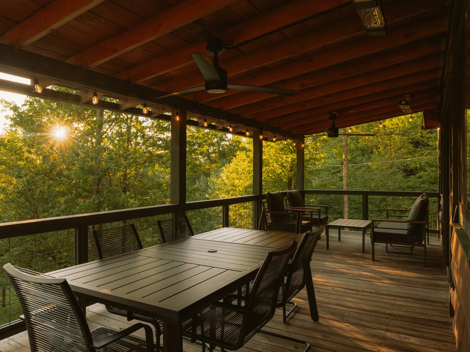 An attached covered deck with timber beams, dark seating and dining furniture, wire railings and a wooded sunset view.
